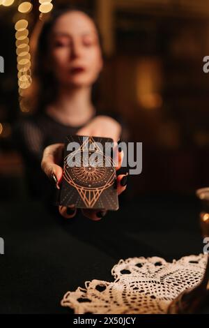 Vertical selective focus shot of mysterious fortuneteller asking client to move deck of cards for layout during session, in dark esoteric room by Stock Photo