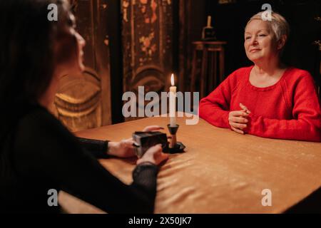 Rear view of enigmatic fortune teller reading tarot cards to mature adult woman client during spiritual ceremony in dark room by light of burning Stock Photo