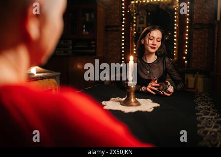 View from shoulder of enigmatic fortune teller reading tarot cards to middle-aged woman client during spiritual ceremony in dark room by light of Stock Photo
