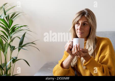 Mature woman in yellow sweater holding a white mug, thoughtfully looking away, with a houseplant beside her Stock Photo