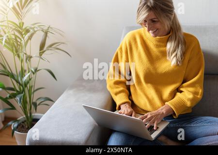 Joyful woman in yellow sweater typing on laptop while sitting on sofa, with a houseplant nearby Stock Photo