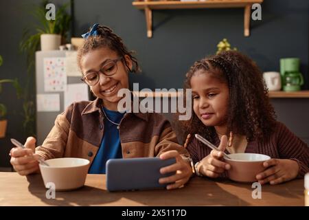 Front view portrait of two smiling African American girls using smartphone at kitchen table in morning scene lit by sunlight Stock Photo