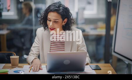 Happy Latin Project Manager with Dark Wavy Hair Working on Laptop in Busy Creative Office. Beautiful Diverse Female Ad Manager Comparing Reports and Graphs on Paper and on Her Computer. Stock Photo