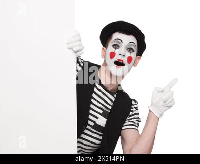 Funny mime artist peeking out of blank poster and pointing at something on white background Stock Photo