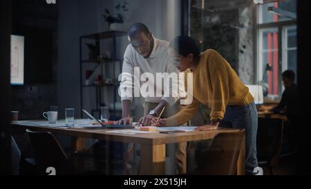 Two Diverse Multiethnic Colleagues Have a Conversation in a Meeting Room Behind Glass Walls in an Agency. African American Creative Director and Female Project Manager Discuss Work on Laptop Computer. Stock Photo