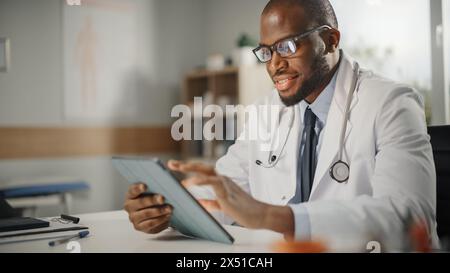 Happy and Smiling African American Male Doctor Wearing White Coat Working on Tablet Computer at His Office. Medical Health Care Professional Working with Test Results, Patient Treatment Planning. Stock Photo