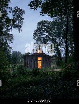 The Apiary pavilion seen at dusk, with surrounding trees silhouetted against the sky. interior lights are golden showing atrium  window. The Beezantiu Stock Photo