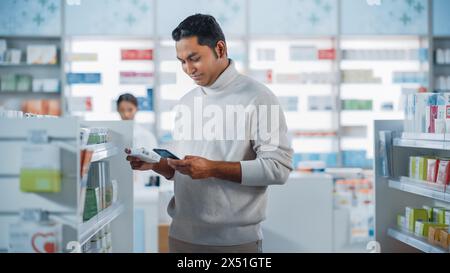 Pharmacy Drugstore: Portrait of a Handsome Young Indian Man Using Smartphone Device, Chooses to Purchase Best Medicine, Drugs, Vitamins. Shelves full of Sport Supplements, Health Care Products Stock Photo