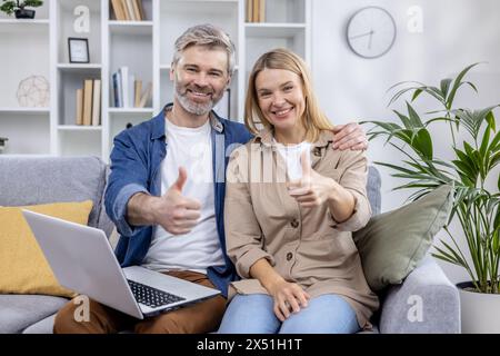 A cheerful middle-aged couple sitting on a couch in their living room, giving thumbs up while using a laptop, surrounded by modern decor. Stock Photo