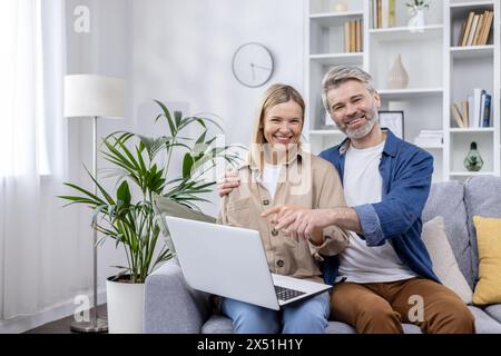 Cheerful mature couple sitting together on the sofa, using a laptop in a well-decorated living room with a friendly and comfortable vibe. Stock Photo