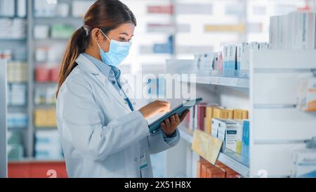 Pharmacy Drugstore: Portrait of Beautiful Pharmacist Wearing Face Mask Uses Digital Tablet Computer, Looks at Camera and Smiles Charmingly, Behind Her Shelves Full of Medicine Packages. Side View Shot Stock Photo