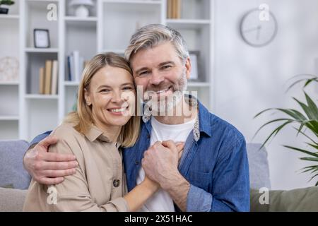 A cheerful middle-aged couple hugs warmly in their stylish living room, smiling at the camera with a bookshelf and modern decor in the background. Stock Photo
