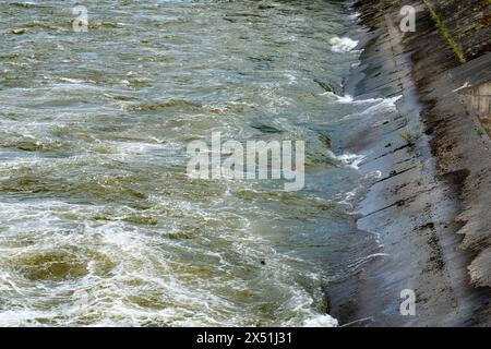 Texture of water with spray rolling against a river bank with weathered and mossy concrete slabs. Stock Photo