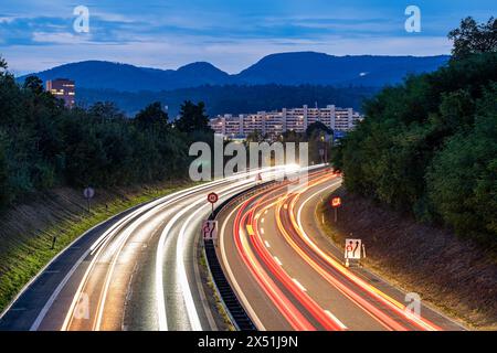 Traffic on a road painting light trails. Road signs warn of a construction site ahead. Aerial view from a bridge. Apartment complex, hills and clouds Stock Photo