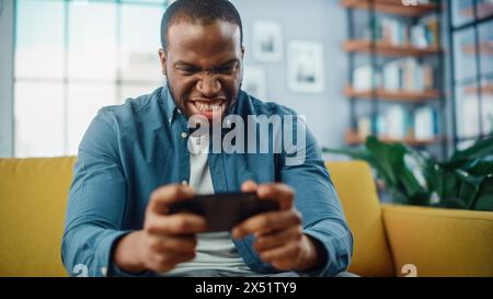 Angry Black African American Man Playing Video Game on Smartphone App while Sitting on a Sofa in Living Room. Excited Person of Color Resting at Home and Having Fun Over the Internet. Stock Photo