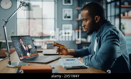 African American Man Having an Online Video Lesson on Laptop Computer while Sitting Behind Desk in Living Room. Freelancer Working on His Skills or Student Doing Homework Over the Internet. Stock Photo