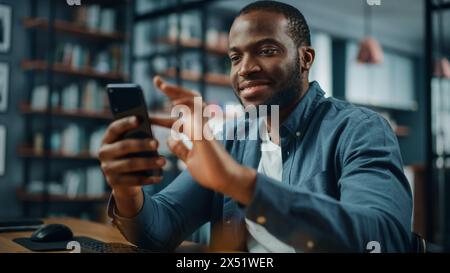 Excited Black African American Man Using Smartphone while Sitting at Table in Living Room. Happy Man Smiling at Home and Chatting to Colleagues and Clients Over the Internet. Using Social Networks. Stock Photo