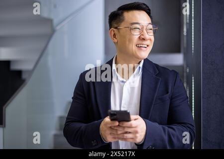 A professional Asian businessman smiling joyfully while using his mobile phone in the contemporary settings of an office staircase, projecting success and connectivity. Stock Photo