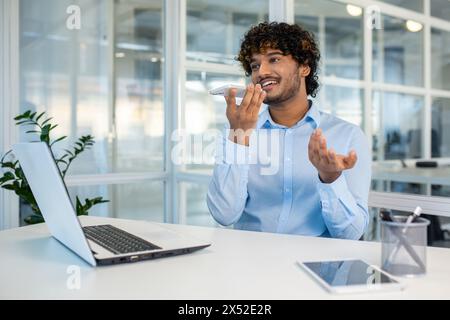 A cheerful young man with curly hair talks animatedly on his smartphone in a modern, well-lit office environment. He appears engaged and happy as he communicates. Stock Photo