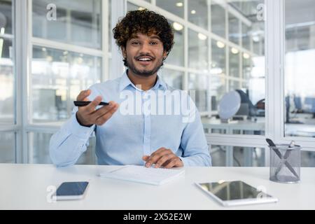 Confident young man with curly hair presents while engaging with colleagues in a well-lit, contemporary office setting. He's holding a remote and smiling at the camera. Stock Photo