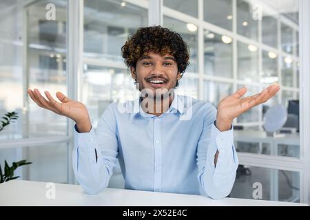 A cheerful young man with curly hair smiling and gesturing in an office environment, looking camera, giving a relaxed and friendly vibe. Stock Photo