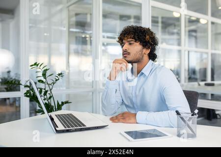 A thoughtful young man with curly hair is sitting at a modern office desk, contemplating while looking away. Bright interior with natural light and green plants. Stock Photo
