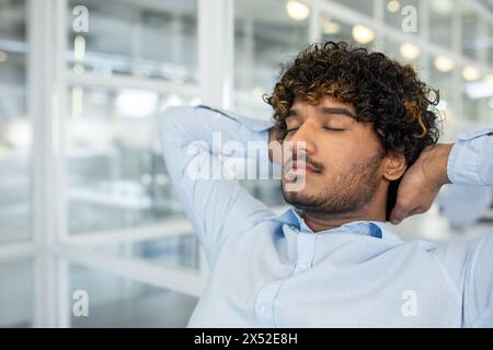 A young man in a light blue button-down shirt relaxes with eyes closed, stretching his neck while seated in a bright modern office space, displaying feelings of ease and peace. Stock Photo