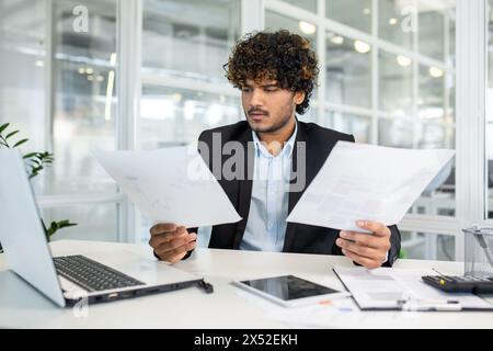 Focused young businessman with curly hair wearing a suit examining multiple documents and reports while sitting at a modern office desk. Stock Photo