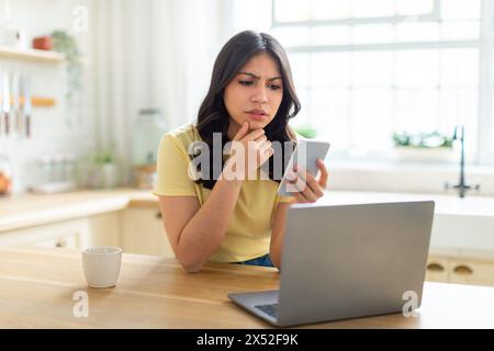 Pensive Young Woman Using Smartphone While Working on Laptop Stock Photo
