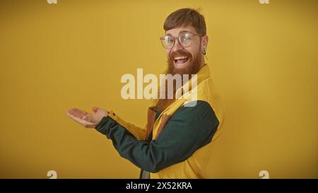 A cheerful bearded man in casual attire presents with an open hand against a solid yellow background Stock Photo