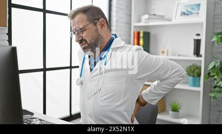 A mature hispanic man in a clinic suffering from back pain while wearing a lab coat and glasses Stock Photo