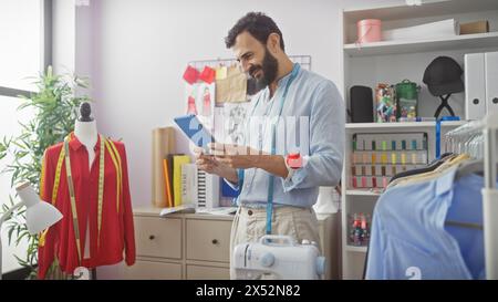 Bearded man using tablet in a bright tailor shop with mannequin, colorful threads, sewing machine, and garments. Stock Photo