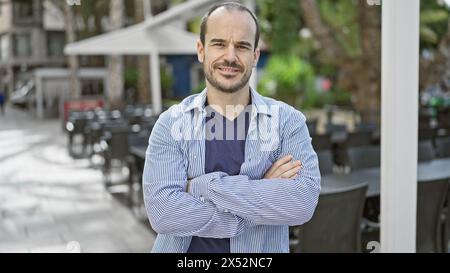 A bearded, bald hispanic man crosses his arms while standing on a city street with an outdoor cafe in the background Stock Photo