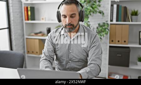 Mature hispanic man with grey beard in headset focusing on laptop in a modern office environment Stock Photo