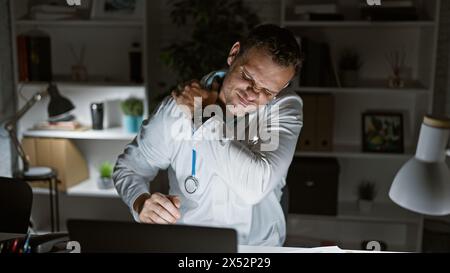 Hispanic male doctor feeling neck pain during a late night at the office, portraying workplace stress. Stock Photo