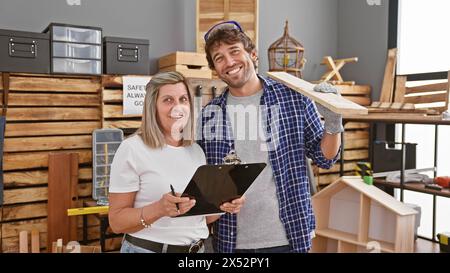 Two smiling carpenters, man and woman, confidently taking notes on clipboard while holding wood plank together in professional indoor carpentry worksh Stock Photo