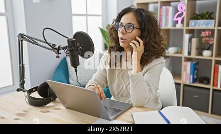 A middle-aged woman talks on the phone while working in a radio studio, surrounded by broadcast equipment. Stock Photo