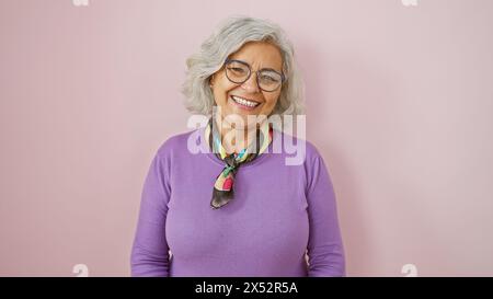 A cheerful mature woman with grey hair, glasses, and wearing a purple sweater, poses against a pink background Stock Photo