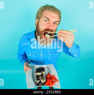 Traditional japanese food. Handsome man in denim shirt eating sushi roll with chopsticks. Sea food. Stylish bearded man with plate of delicious maki Stock Photo