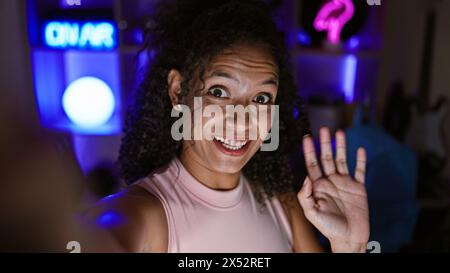 Confident curly-haired young hispanic woman streamer rocks her gaming with brilliant smile during late-night video call in gaming room. Stock Photo