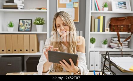 Blonde woman wearing white sweater using tablet in modern office interior with shelves and decorations. Stock Photo