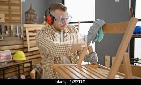 Bearded man in safety gear meticulously cleaning a wooden chair in a well-organized carpentry workshop. Stock Photo