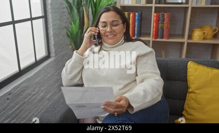 A mature hispanic woman smiling while talking on the phone and holding documents in a cozy living room. Stock Photo