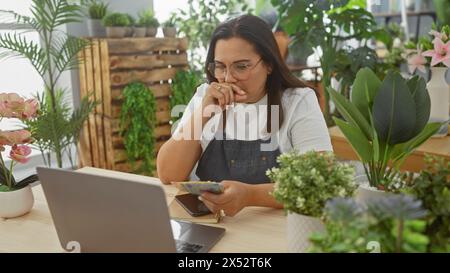 Middle-aged hispanic woman accounting in a green flower shop with laptop and lei banknotes. Stock Photo