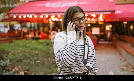 A brunette woman talks on her smartphone at an outdoor cafe terrace during evening hours. Stock Photo
