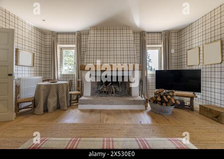 A large living room with a fireplace with wooden floors, natural fiber carpet, flat TV, a basket with firewood and square windows Stock Photo