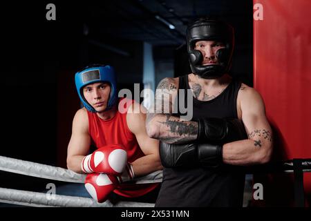 Two young boxers in black and red sportswear and protective sports helmets or shields standing on boxing ring and looking at camera Stock Photo