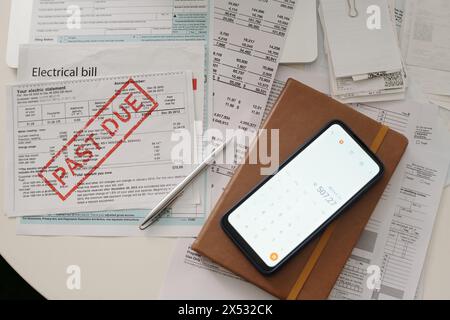 Sum of unpaid financial paper on screen of smartphone lying on top of notebook in leather cover and electrical bill with past due stamp Stock Photo