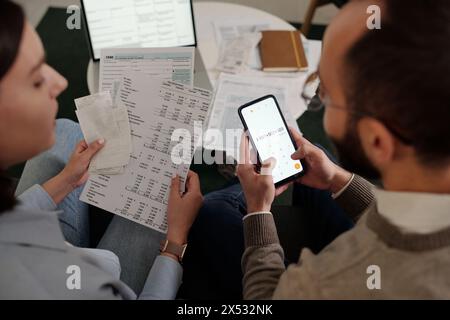 High angle of young couple sitting by table and discussing sums in financial documents while man with smartphone making calculations Stock Photo