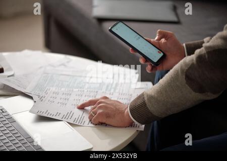 Hand of young unrecognizable man pointing at financial bill with several columns of sums and using calculator in mobile phone Stock Photo